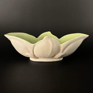 May include: A cream-colored ceramic bowl with a light green interior. The bowl is shaped like a stylized flower with curved petals and a rounded base. The bowl is on a black background.