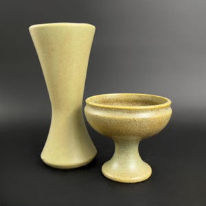 May include: Two ceramic vases in a speckled light green glaze. The taller vase is a slender cylinder with a flared top. The shorter vase is a bowl on a pedestal.
