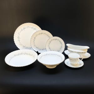 Może przedstawiać: Zestaw białej porcelanowej zastawy stołowej z delikatnym niebieskim kwiatowym wzorem. Zestaw zawiera talerze o różnych rozmiarach, miskę do serwowania, sosjerkę i filiżankę do herbaty ze spodkiem. Krawędzie talerzy są faliste.