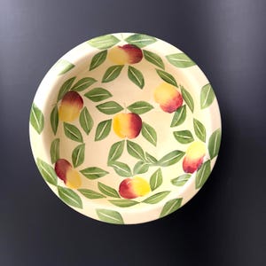 May include: A round, cream-colored ceramic bowl with a hand-painted design of apples and green leaves. The apples are red and yellow, and the leaves are green. The bowl is set against a dark background.