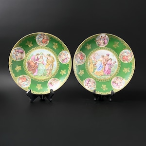 May include: Two decorative plates with a green background and gold trim. Each plate features a central image of people in a garden setting, surrounded by smaller images of people and cherubs.
