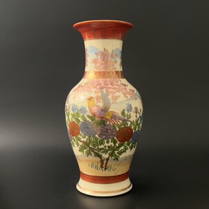 May include: A white ceramic vase with a red rim and base. The vase is decorated with a colorful floral and bird design. The flowers are pink, blue, and red, and the bird is yellow, red, and green.