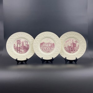 May include: Three white ceramic plates with raised edges and pink images of buildings. The plates are on black stands.