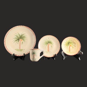 May include: A cream-colored ceramic dinnerware set featuring a palm tree design. The set includes a large plate, a smaller plate, a bowl, and a mug. The plates have a red and white patterned border. The palm trees are green with a brown trunk.