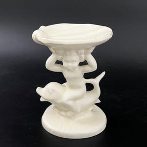May include: White ceramic trinket dish featuring a cherub riding a dolphin and holding a shell above its head.