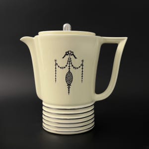 Könnte beinhalten: Eine weiße Kaffeekanne aus Keramik mit einem silbernen Deckel und schwarzen und silbernen Dekordetails. Die Kanne hat einen runden Korpus mit einem ausgestellten Ausguss und einem Henkel.