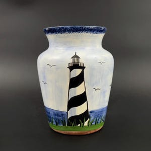 May include: A white ceramic vase with a blue rim and a hand-painted design of a black and white striped lighthouse against a blue sky with white clouds and seagulls. The lighthouse is set against a blue ocean with green grass.