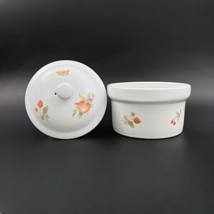 May include: White ceramic bowl with a lid, both decorated with a floral pattern featuring strawberries, cherries, and a butterfly. The bowl has a round shape and a slightly raised rim.