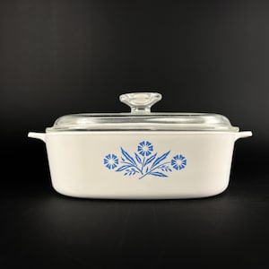 Op de afbeelding: Een witte rechthoekige ovenschaal met een doorzichtig glazen deksel. De schaal heeft een blauw bloemenontwerp. De deksel heeft een doorzichtige glazen knop. De schaal heeft twee handvatten.
