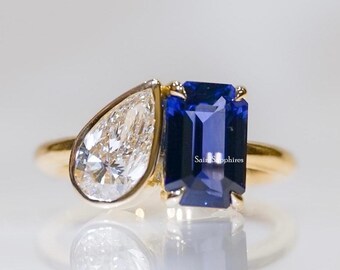 May include: A gold ring featuring two prominent stones: a pear-shaped clear diamond and a rectangular emerald-cut blue sapphire. The stones are set side-by-side on a polished gold band, creating a distinct two-stone design with clear reflections.