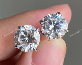 May include: A pair of round-cut diamond stud earrings. The earrings feature large, sparkling, clear gemstones set in a four-prong setting. The earrings are displayed against a neutral background.