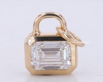 May include: A gold-toned pendant shaped like a miniature padlock, featuring a rectangular emerald-cut clear gemstone set in the center. The piece includes a small attached jump ring at the top for hanging, with a polished metallic finish.