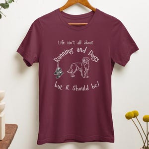 以下が含まれることがあります： 白のテキストとグラフィックが入ったバーガンディのTシャツ。「Life isn't all about Running and Dogs but it should be!」というテキスト。犬とランニングシューズがテキストの下に描かれています。シャツは木製のハンガーにかかっています。