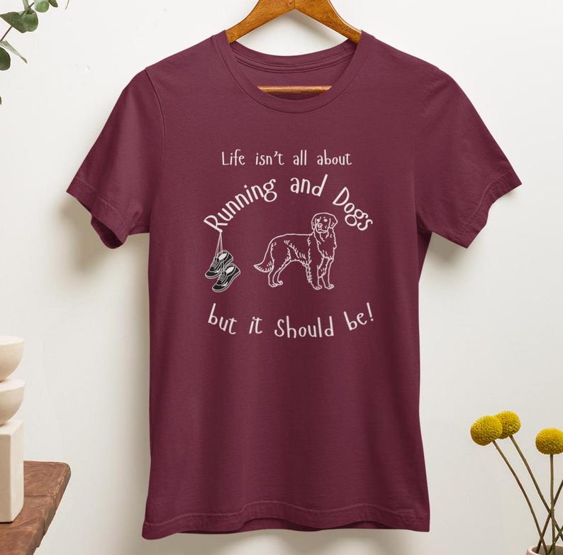 以下が含まれることがあります： 白のテキストとグラフィックが入ったバーガンディのTシャツ。「Life isn't all about Running and Dogs but it should be!」というテキスト。犬とランニングシューズがテキストの下に描かれています。シャツは木製のハンガーにかかっています。