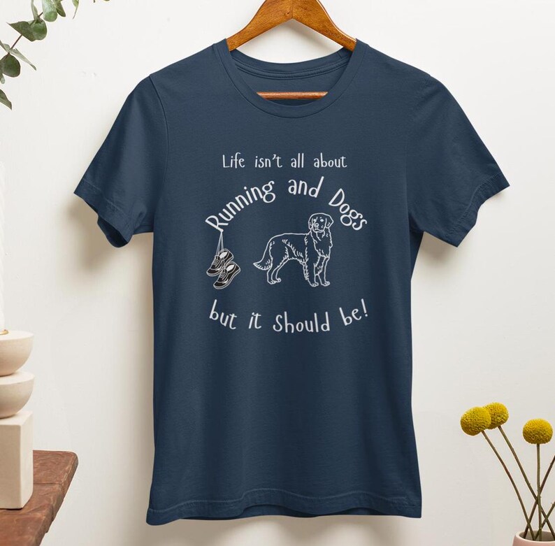 以下が含まれることがあります： 白のテキストとグラフィックが特徴のネイビーブルーのTシャツ。「Life isn't all about Running and Dogs but it should be!」というテキストと犬のイラスト、ランニングシューズが描かれています。