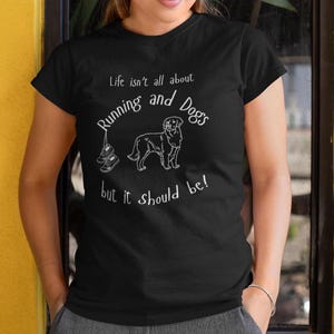 以下が含まれることがあります： 白のテキストとグラフィックが入った黒いTシャツ。「人生はランニングと犬だけではありませんが、そうあるべきです！」というテキスト。犬とランニングシューズの線画も特徴です。