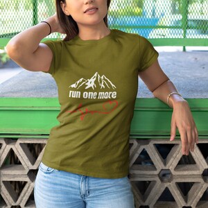 May include: A woman wearing a green t-shirt with a white mountain graphic and the text "run one more" with a red heart beat line.