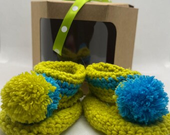 May include: Handmade crochet baby booties in bright yellow with blue accents and pom-poms. The booties are presented in a small brown gift box with a clear window and a green polka dot ribbon. A charming and colorful baby gift.