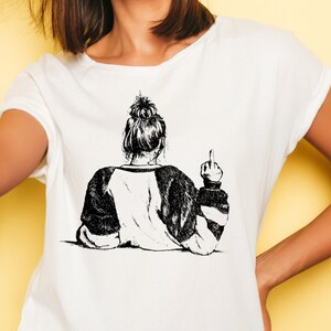 May include: A white t-shirt featuring a black and white illustration of a woman with her hair in a bun giving the middle finger.