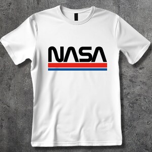Może przedstawiać: Biała koszulka z czarnym logo NASA i czerwonymi i niebieskimi paskami.