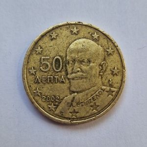 May include: A gold-colored Greek coin with a portrait of Eleftherios Venizelos and the text "50 ΛΕΠΤΑ" and "ΕΛ. ΒΕΝΙΖΕΛΟΣ". The coin also has the year "2002" and 12 stars around the edge.