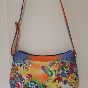May include: A colorful leather handbag with a floral and hummingbird design. The bag has a long strap and a zipper closure.
