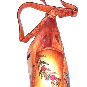 May include: A brown leather shoulder bag with a floral design. The bag has a long strap and a flap closure. The flowers are painted in shades of red, blue, and green.