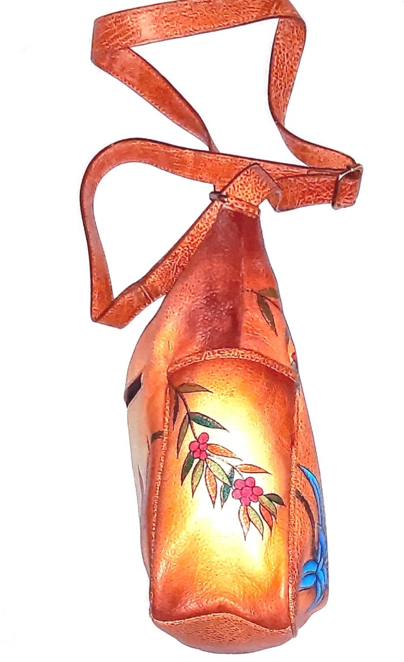 May include: A brown leather shoulder bag with a floral design. The bag has a long strap and a flap closure. The flowers are painted in shades of red, blue, and green.