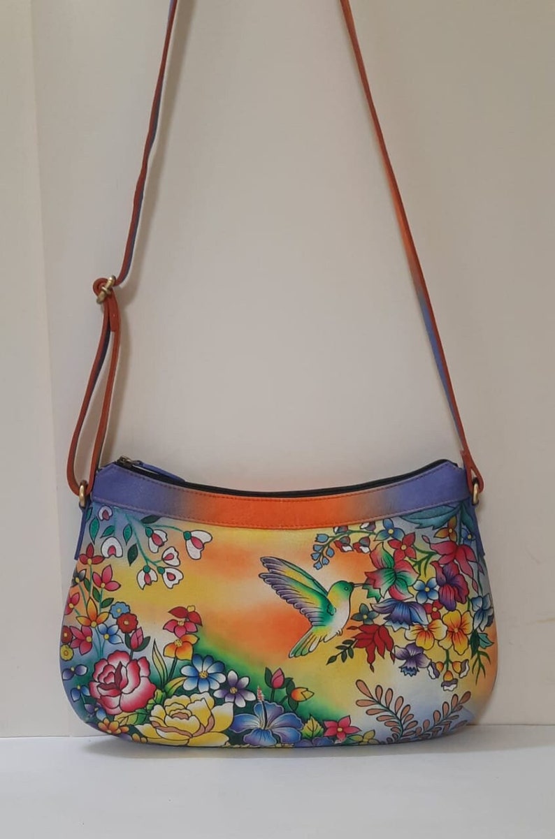 May include: A colorful leather handbag with a floral and hummingbird design. The bag has a long strap and a zipper closure.