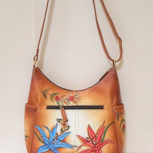 May include: A brown leather shoulder bag with a floral design. The bag features two large flowers, one blue and one red, with a butterfly in the center. The bag has a zippered pocket on the front and a strap that can be adjusted.