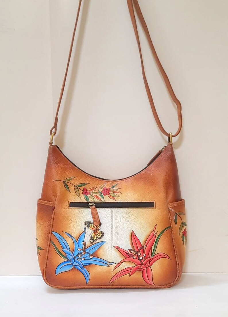 May include: A brown leather shoulder bag with a floral design. The bag features two large flowers, one blue and one red, with a butterfly in the center. The bag has a zippered pocket on the front and a strap that can be adjusted.