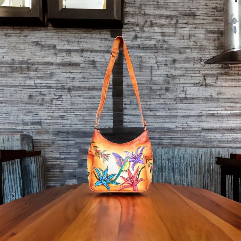 May include: A tan leather shoulder bag with a floral and hummingbird design. The bag has a long strap and a flap closure.