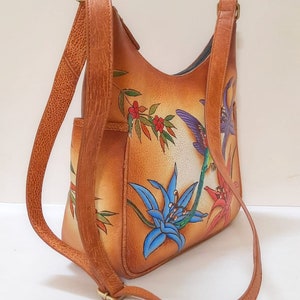 May include: A brown leather shoulder bag with a floral design featuring hummingbirds and lilies. The bag has a long adjustable strap.