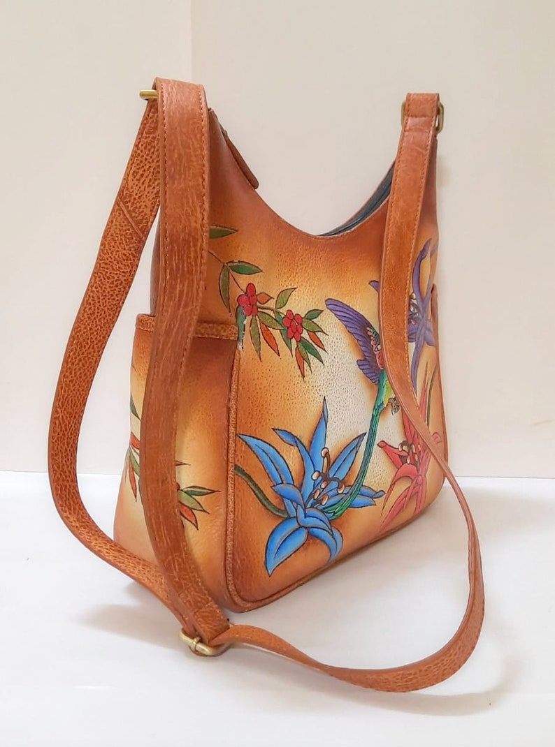 May include: A brown leather shoulder bag with a floral design featuring hummingbirds and lilies. The bag has a long adjustable strap.