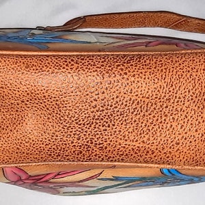 May include: A brown leather handbag with a floral design. The bag has a top handle and a shoulder strap. The flowers are painted in shades of pink, blue, and green.