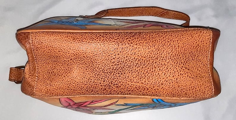May include: A brown leather handbag with a floral design. The bag has a top handle and a shoulder strap. The flowers are painted in shades of pink, blue, and green.