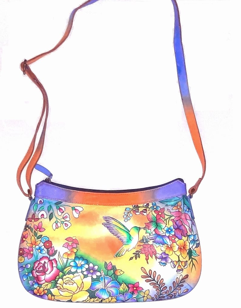 May include: A colorful leather handbag with a hummingbird and floral design. The bag has a long strap and a flap closure.
