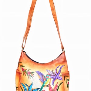 May include: A tan leather shoulder bag with a painted design of hummingbirds and flowers. The bag has a front pocket and a long adjustable strap.