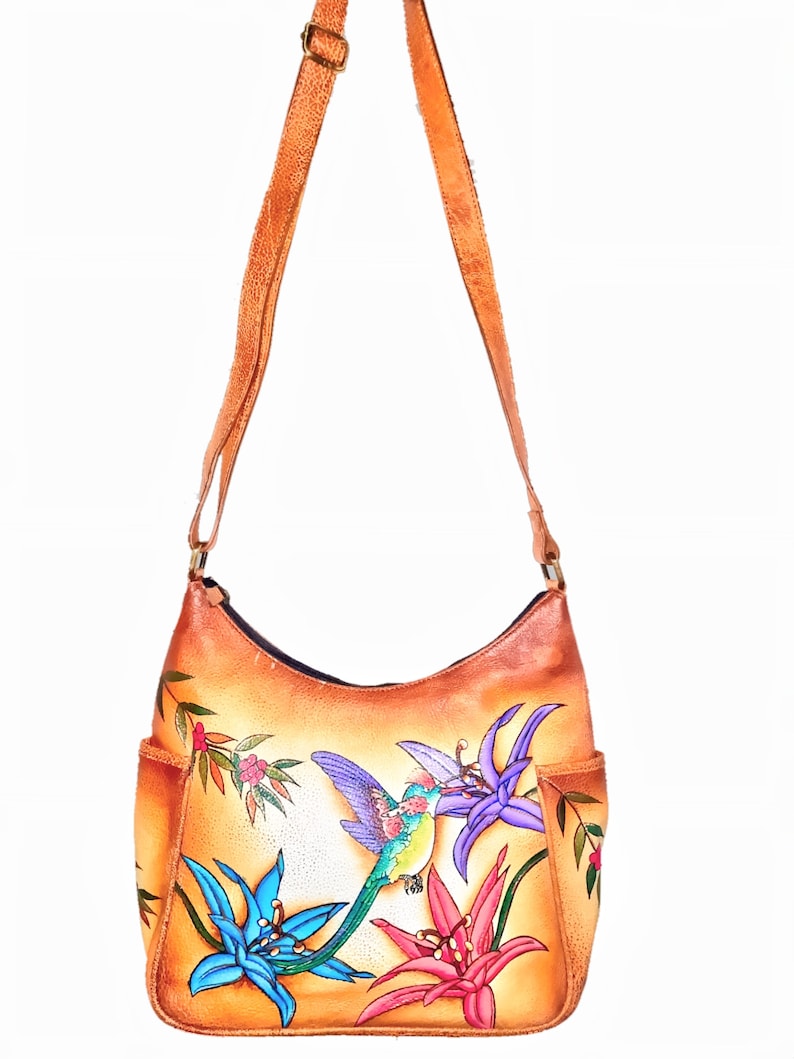 May include: A tan leather shoulder bag with a painted design of hummingbirds and flowers. The bag has a front pocket and a long adjustable strap.