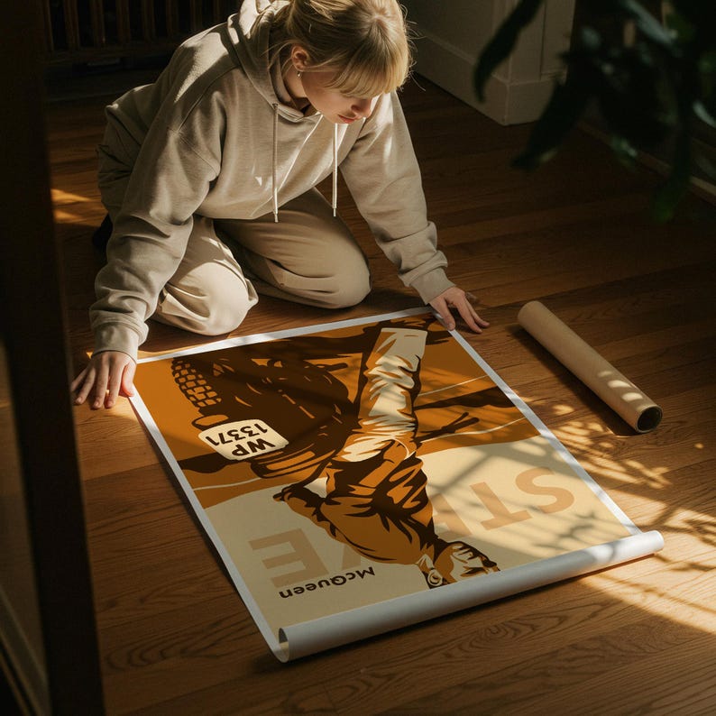 May include: A rolled poster featuring a vintage-style illustration in shades of brown and beige. The poster includes the text "McQueen" and "WP 13371". The poster is being unrolled on a wooden floor.