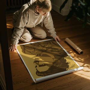 May include: A rolled poster featuring a sepia-toned illustration of a person's profile. The artwork has a minimalist style with bold lines and a solid background. The poster is unrolled on a wooden floor.