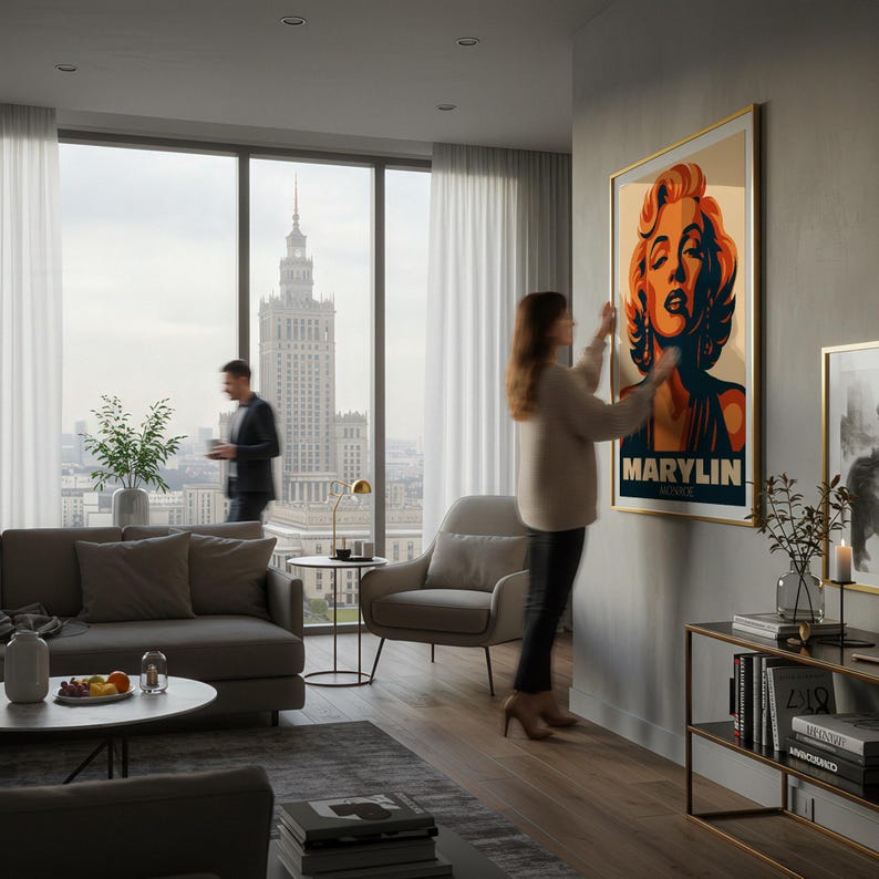 May include: A framed art print featuring a stylized portrait of Marilyn Monroe with the name "MARYLIN" at the bottom. The artwork has a gold frame and is being hung on a gray wall in a modern living room. The room has large windows with a city view.