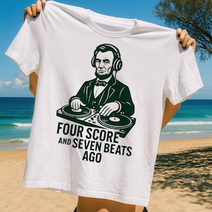 Może przedstawiać: Biała koszulka z zieloną grafiką Abrahama Lincolna w słuchawkach jako DJ. Pod obrazkiem znajduje się napis "FOUR SCORE AND SEVEN BEATS AGO". Koszulka jest trzymana przez osobę na plaży.