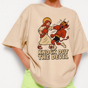 Könnte beinhalten: Beiges T-Shirt mit einer Cartoon-Illustration von Jesus, der den Teufel boxt. Jesus trägt ein Gewand und Boxhandschuhe, während der Teufel rot mit Hörnern und Boxshorts ist. Der Text "KNOCK OUT THE DEVIL" ist darunter gedruckt.