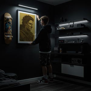 May include: A framed portrait with a gold and brown color scheme is being hung on a dark wall. A skateboard leans against the wall. The room has dark furniture and LED lighting.