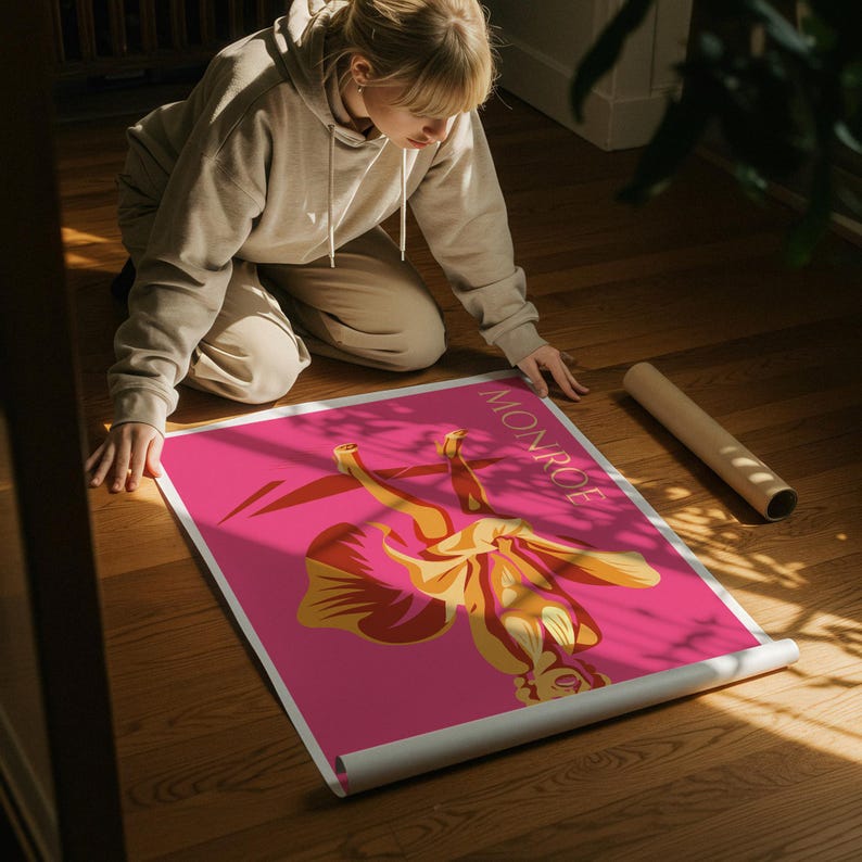 Puede incluir: Un p&oacute;ster enrollado con una ilustraci&oacute;n estilizada de una mujer con un vestido fluido sobre un fondo rosa. El p&oacute;ster tiene el nombre "MONROE" impreso en rosa claro. El p&oacute;ster est&aacute; sobre un suelo de madera.