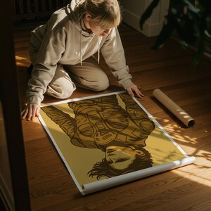 May include: A person kneeling on a wooden floor, unrolling a poster. The poster features a sepia-toned illustration of a person in a futuristic outfit. A rolled-up poster is also on the floor.