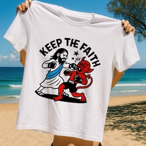 Puede incluir: Camiseta blanca con una ilustración de dibujos animados de Jesús boxeando contra un diablo rojo. El texto "KEEP THE FAITH" está impreso encima de las figuras. El diablo lleva pantalones cortos negros con "MMA" impreso.