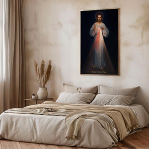 May include: A framed religious artwork depicting a figure in white robes with a red and white light emanating from the chest. The text "Jesus, I trust in You" is at the bottom. The artwork is displayed above a bed with beige bedding.