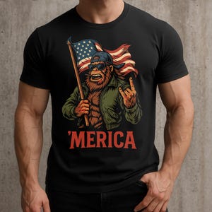 May include: Black t-shirt with a cartoon Sasquatch wearing sunglasses and a baseball cap, holding an American flag. The Sasquatch is giving a rock and roll hand gesture. The word "'MERICA" is printed in red below the graphic.